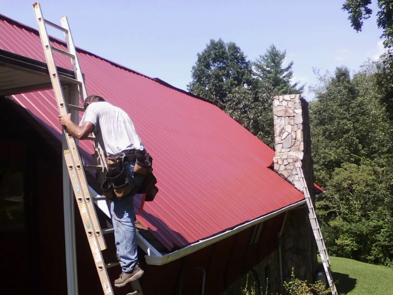 Roofer on a ladder beside a red metal roof and stone chimney — Soffit & Fascia Repair by Roofing Brothers in Canyon Country